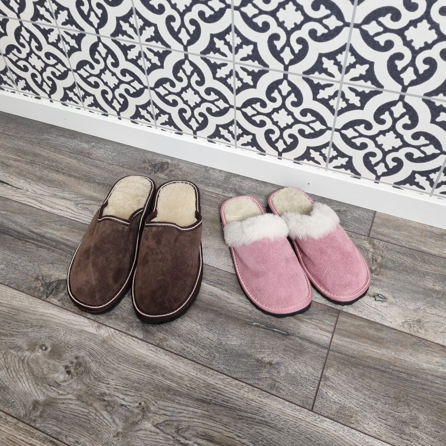 Ett par bruna fårskinnstofflor utan häl och ett par rosa fårskinnstofflor med vit ullkant, placerade på trägolv framför dekorativ svartvit kakelvägg.