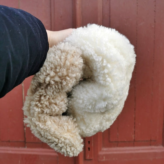 Två fårskinnsadelskydd i ljusbrunt och vitt hålls i handen framför en röd trädörr.