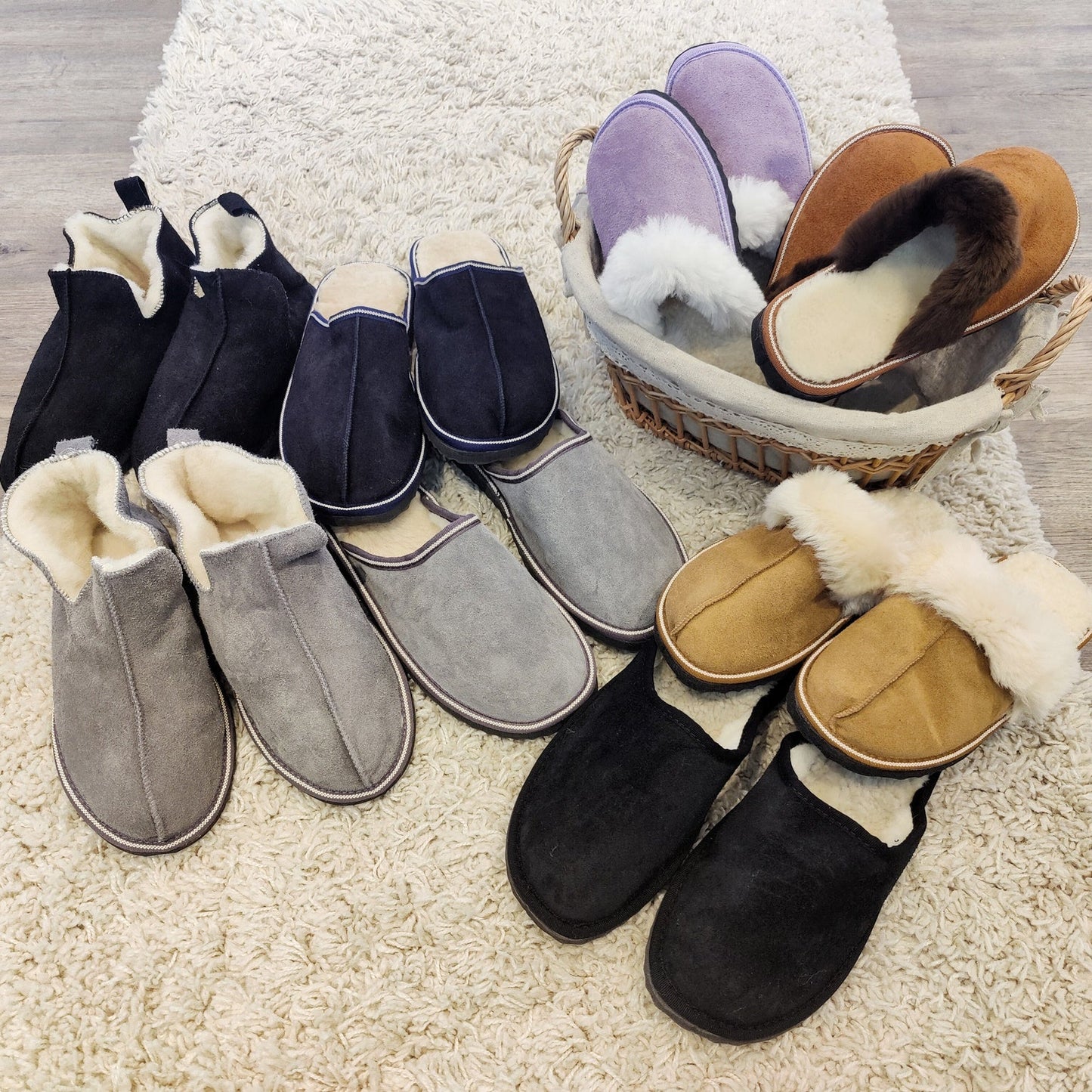 Olika modeller av fårskinnstofflor i flera färger – grå, svart, lila, brun och beige – liggande på matta och i korg.