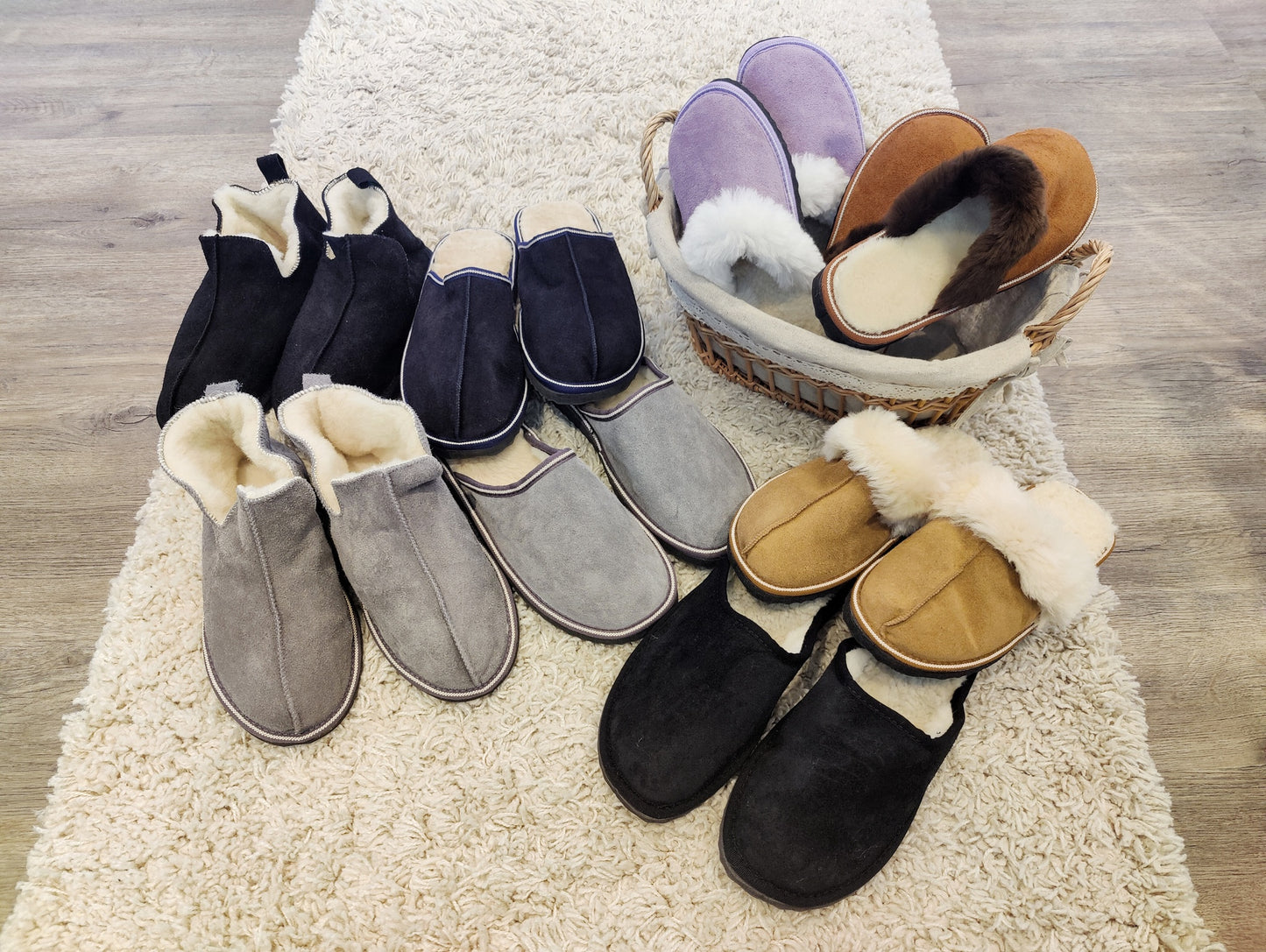 Blandade modeller av fårskinnstofflor och ulltofflor från Jakobs Handmade i olika färger som svart, grå, brun och lila, arrangerade på matta och i korg på trägolv.