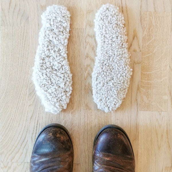 Ett par naturvita fårskinnsulor placerade framför ett par slitna läderskor på trägolv, perfekta för att hålla fötterna varma under vintern.