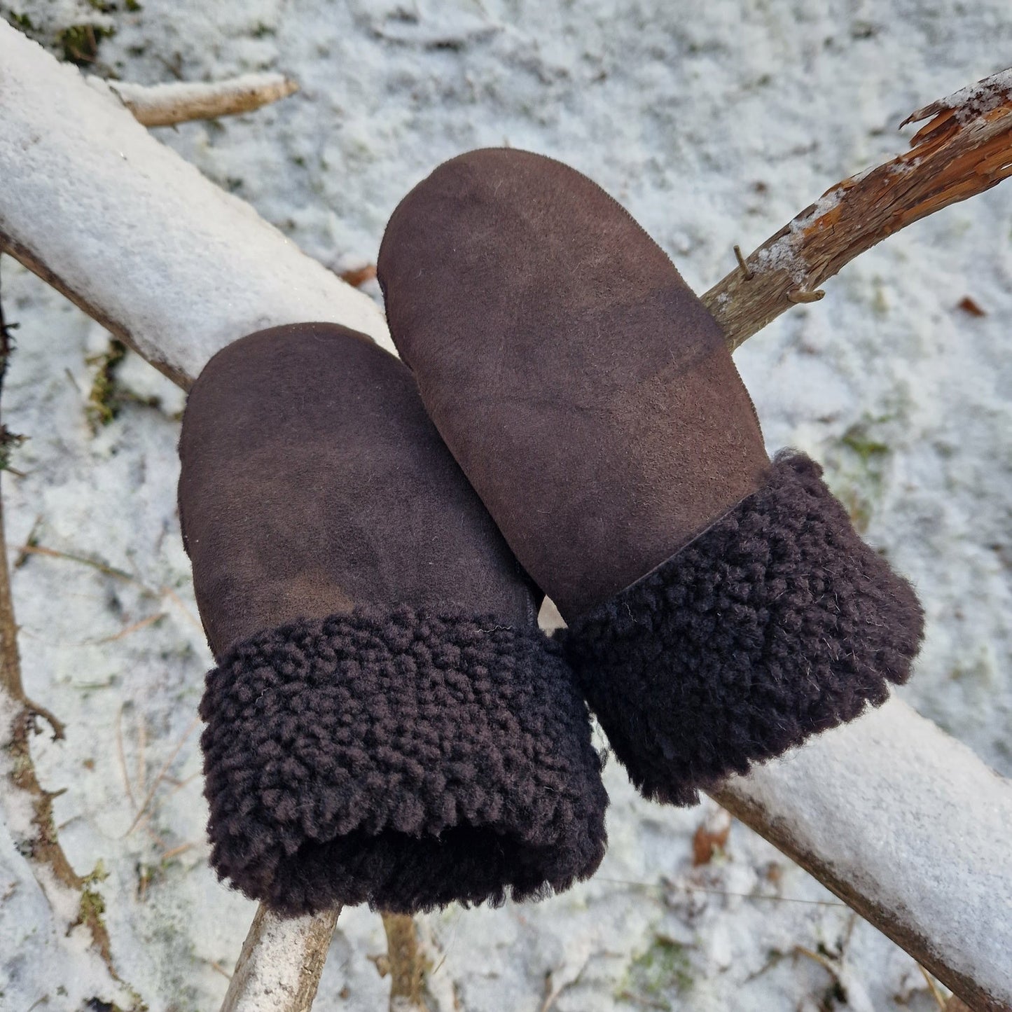 Bruna handgjorda fårskinnsvantar med mörkbrunt ullfoder, liggande på en snötäckt gren – varma och slitstarka vintervantar i äkta fårskinn.