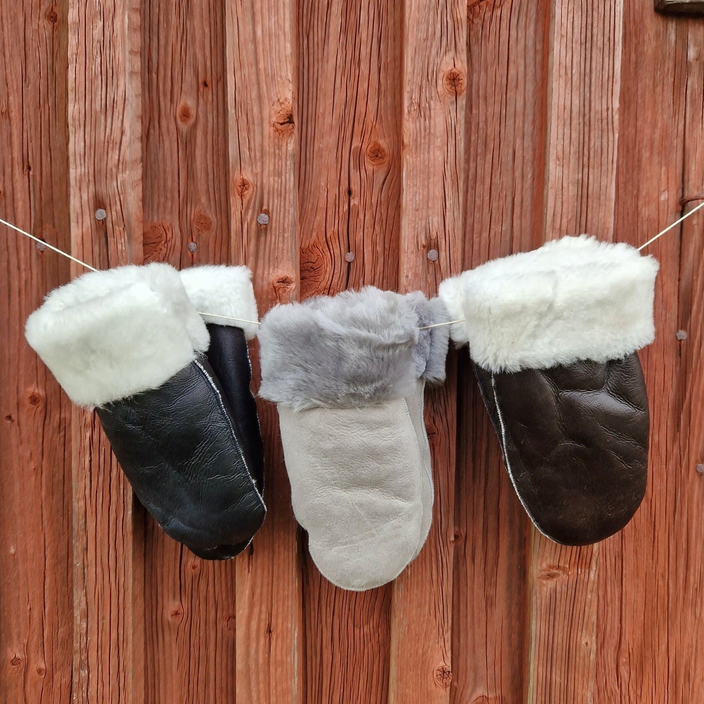 Tre par handgjorda fårskinnsvantar i svart, grå och brun färg, alla med mjukt vitt ullfoder, hänger på en snodd mot en röd trävägg – varma och slitstarka vintervantar för dam och herr.