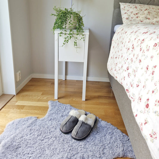 Grå fårskinnstofflor med vitt ullfoder placerade på lila fårskinnsmatta i sovrum, bredvid säng och nattduksbord.