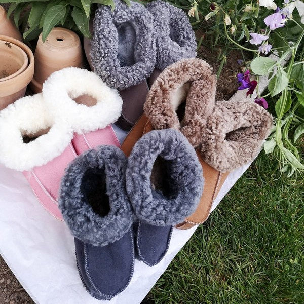 Handgjorda fårskinnstofflor i flera färger från Jakobs Handmade, placerade vid färgglada sommarblommor. Tillverkade i äkta svenskt fårskinn med mjuk, värmande krage.”