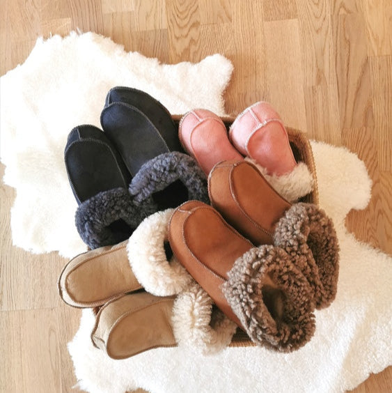 Handgjorda fårskinnstofflor i flera färger från Jakobs Handmade, placerade på ett vitt fårskinn. Tillverkade i äkta svenskt fårskinn med varm och mjuk krage.