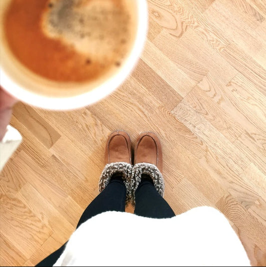 Person i handgjorda ljusbruna fårskinnstofflor från Jakobs Handmade håller en kopp kaffe. Tofflorna är tillverkade i äkta svenskt fårskinn med mjuk brun krage för värme och komfort.
