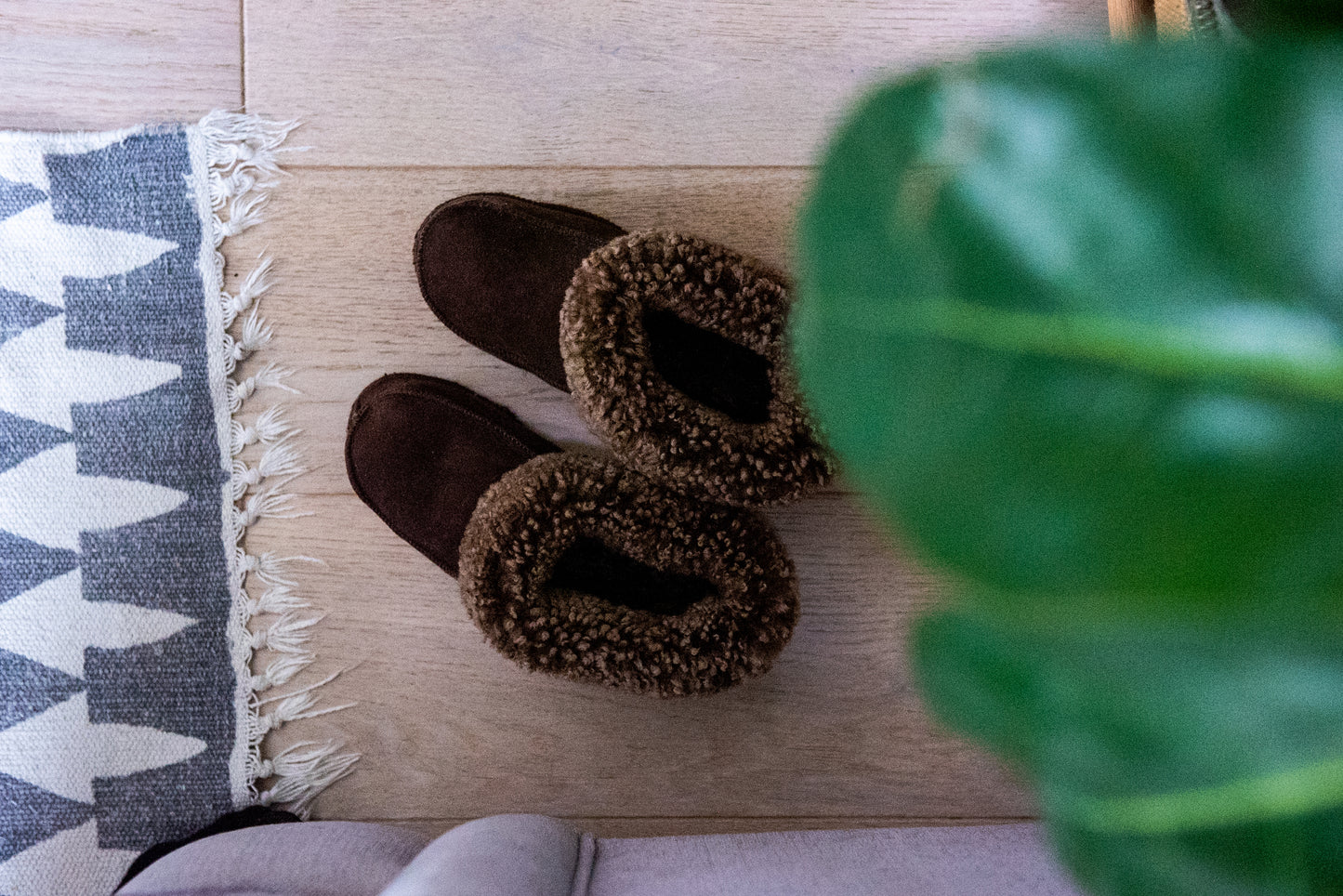 Handgjorda mörkbruna fårskinnstofflor från Jakobs Handmade placerade på ett trägolv bredvid en matta och grön växt. Tillverkade i äkta svenskt fårskinn med mjuk värmande krage.