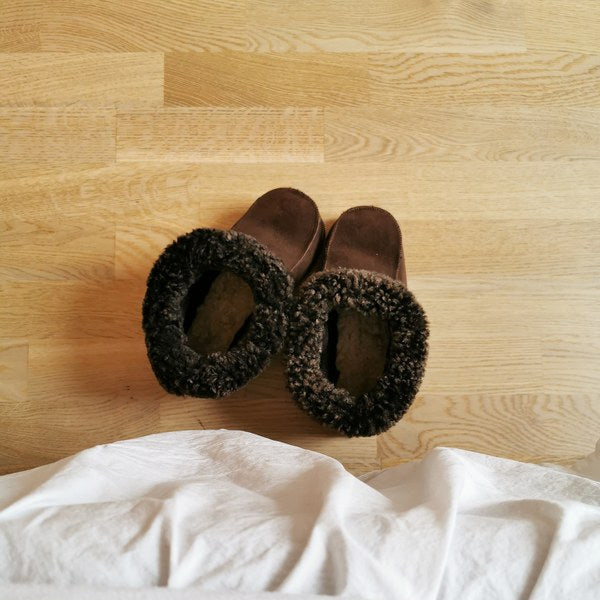 Handgjorda mörkbruna fårskinnstofflor från Jakobs Handmade, placerade på ett trägolv vid en säng. Tillverkade i äkta svenskt fårskinn med mjuk och värmande krage.