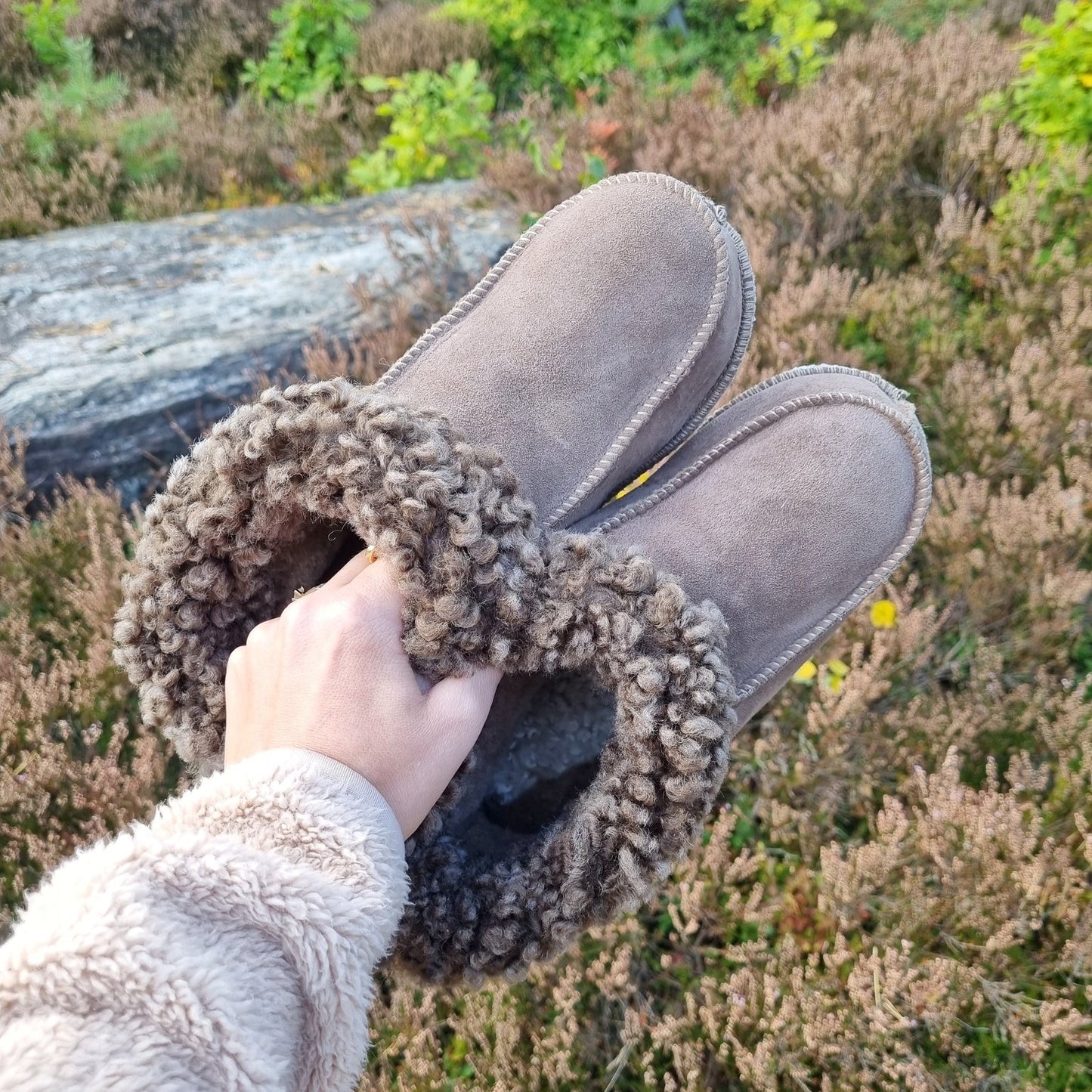 Hand håller ett par handgjorda taupe fårskinnstofflor från Jakobs Handmade, fotograferade i naturen. Tofflorna är tillverkade i äkta svenskt fårskinn med mjuk brun krage.