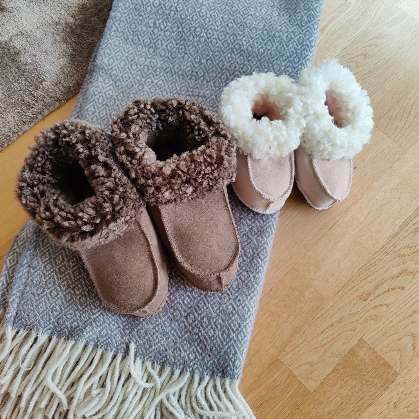 Handgjorda fårskinnstofflor i taupe och rosa från Jakobs Handmade, placerade på en grå ullpläd. Tillverkade i äkta svenskt fårskinn med mjuk värmande krage.