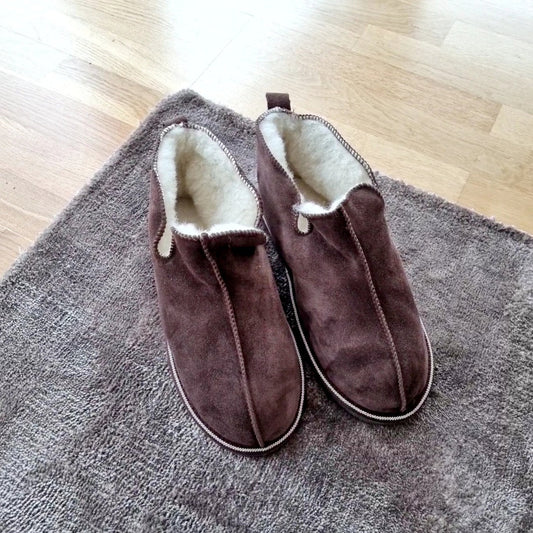 Bruna Kosmos merinoullstofflor från Jakobs Handmade, placerade på en matta – varma och handgjorda tofflor i fårskinn för dam och herr.