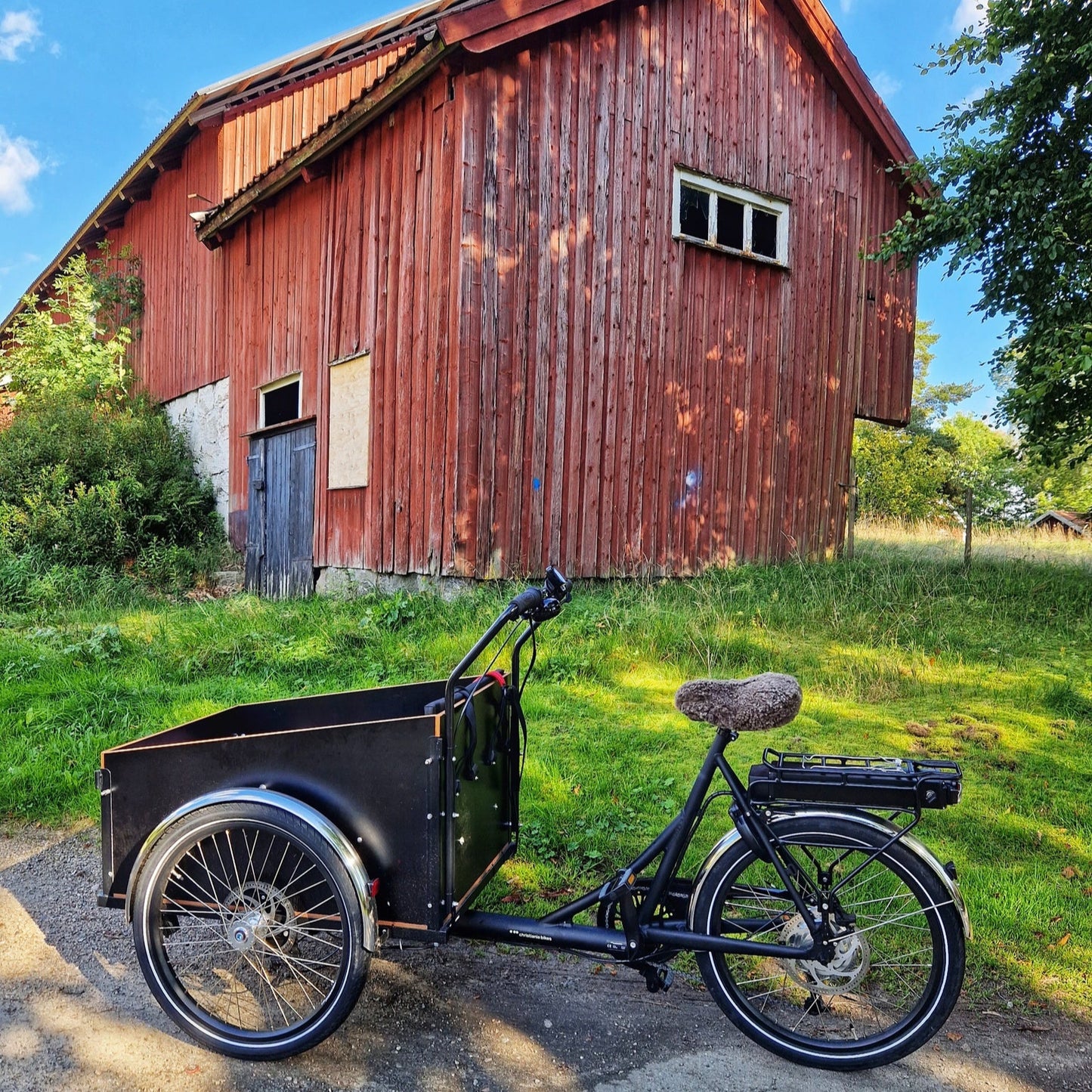 Svart lådcykel med ljusbrunt fårskinnsadelskydd parkerad framför en röd trälada en solig dag.