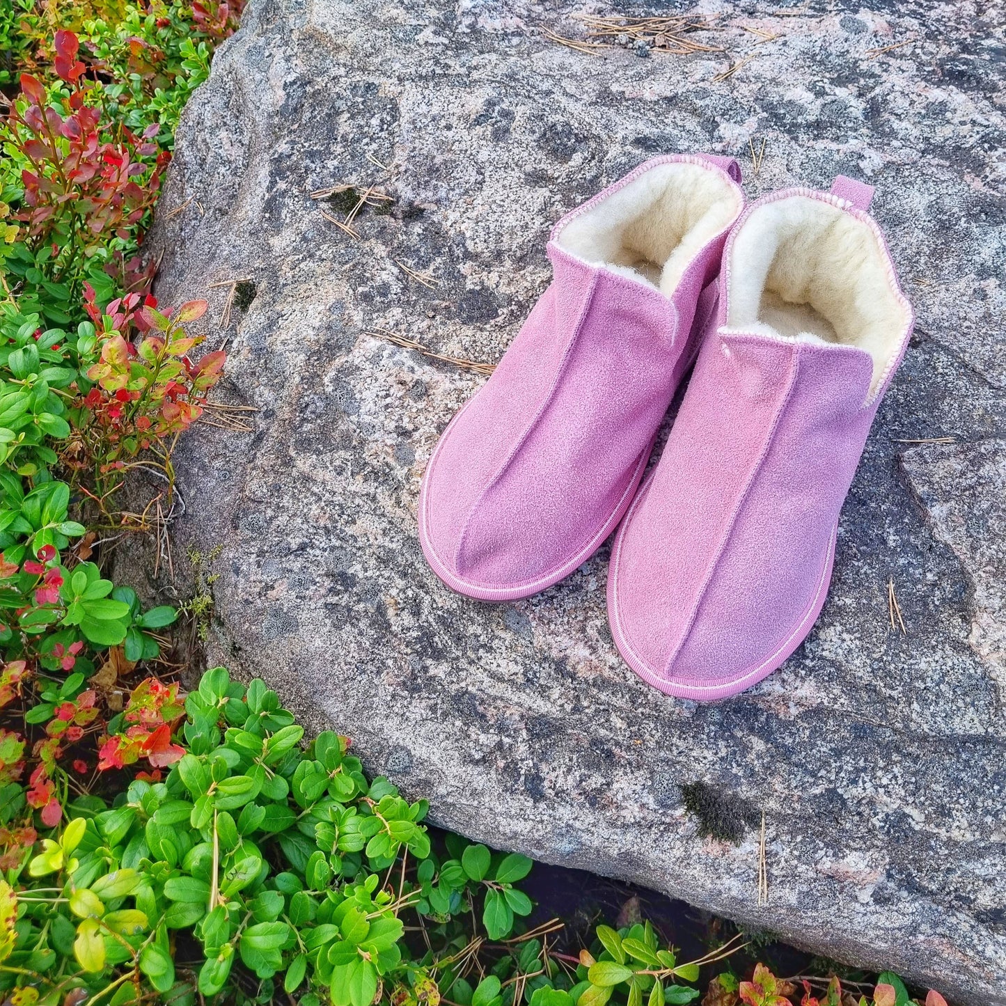 Rosa fårskinnstofflor i ankelmodell med vitt ullfoder för dam, placerade på en sten med höstfärgad växtlighet runtomkring.