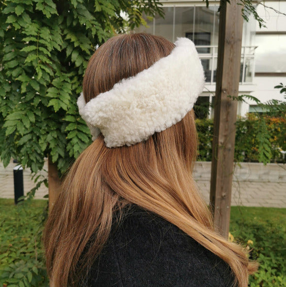 Kvinna med långt brunt hår och svart jacka bär ett vitt fårskinnspannband utomhus framför gr