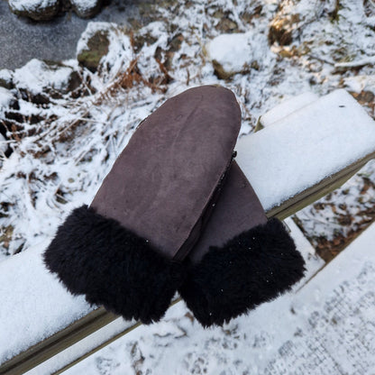 Ett par bruna fårskinnsvantar med svart ullfoder liggande på ett snötäckt räcke i vintrig skogsmiljö – varma och mjuka handgjorda vinterhandskar i äkta fårskinn.