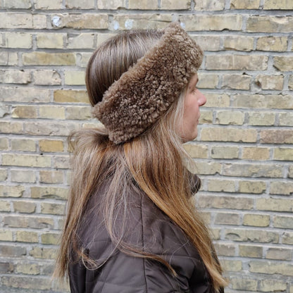 Kvinna med långt brunt hår och brun jacka bär ett brunt fårskinnspannband framför en tegelvägg.