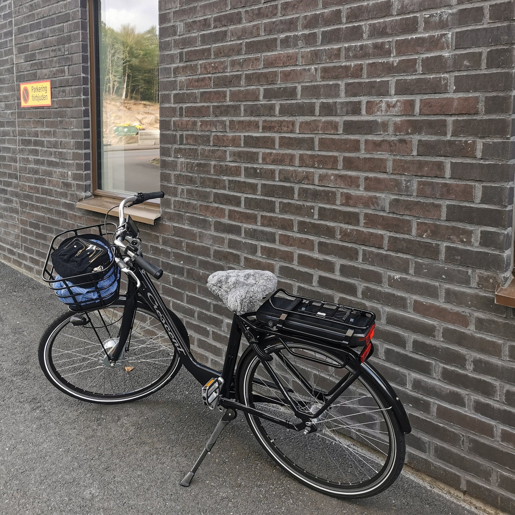 Svart cykel med ljusgrått fårskinnsadelskydd parkerad vid tegelvägg, ger komfort och värme vid cykling.