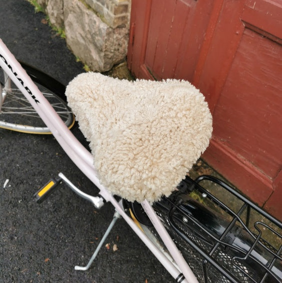 Ljusrosa cykel med ljust fårskinnsadelskydd parkerad framför en röd dörr och tegelvägg.