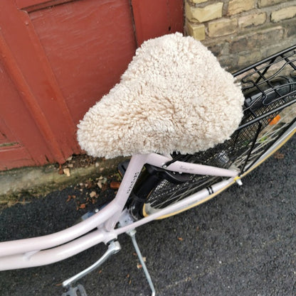 Ljusrosa cykel med ljust fårskinnsadelskydd parkerad framför en röd dörr och tegelvägg.