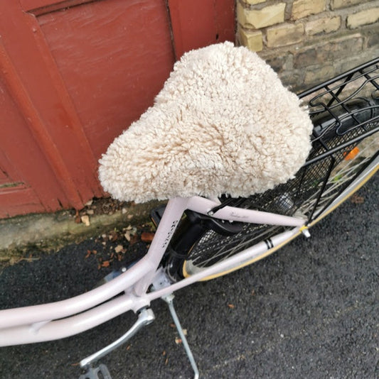 Ljusrosa cykel med ljust fårskinnsadelskydd parkerad framför en röd dörr och tegelvägg.