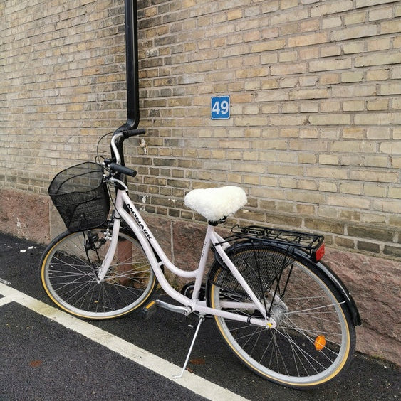 Vit damcykel med vitt fårskinnsadelskydd parkerad vid tegelvägg med husnummer 49.