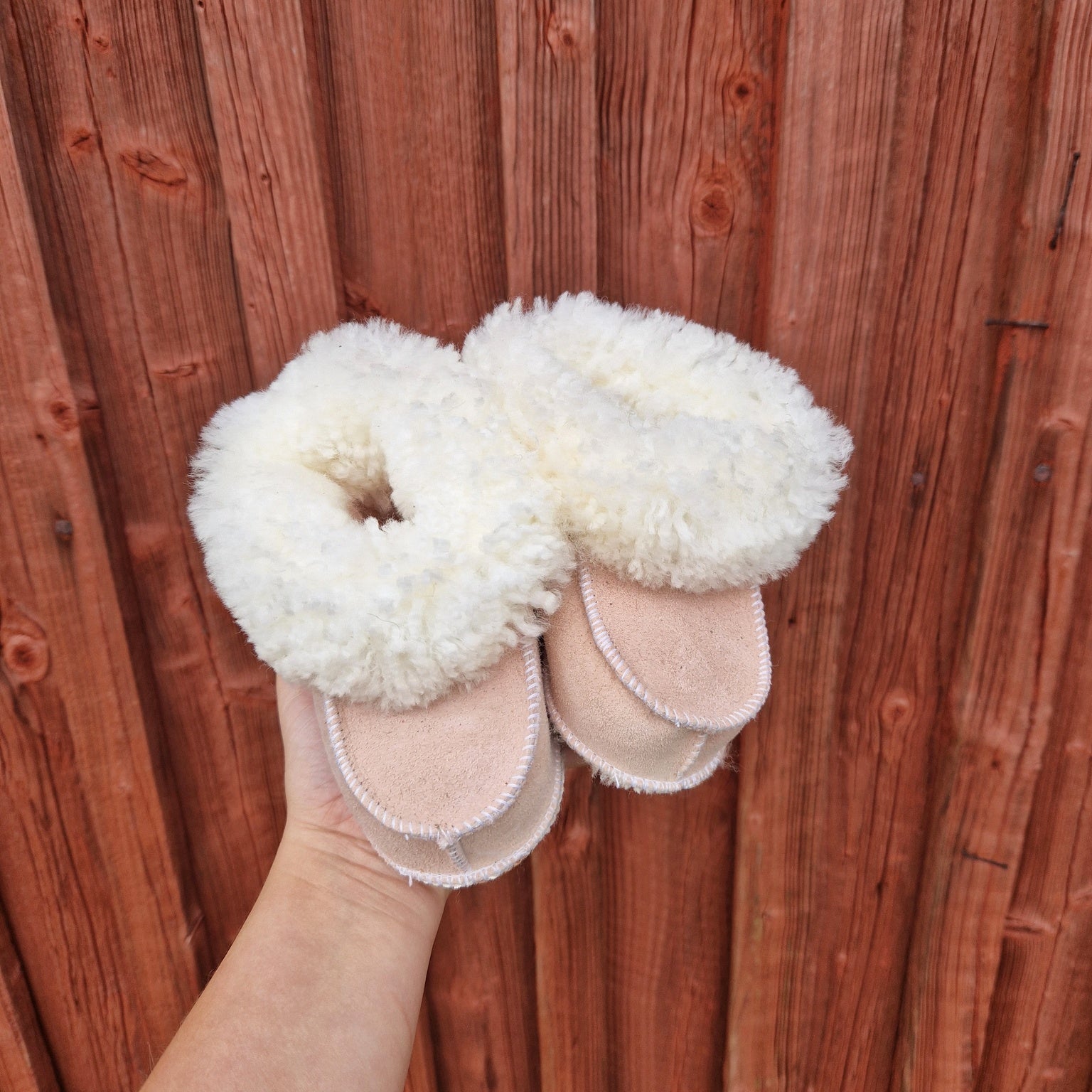 Handgjorda rosa fårskinnstofflor för barn från Jakobs Handmade, i äkta svenskt fårskinn med vit fluffig krage, fotograferade mot en trävägg.