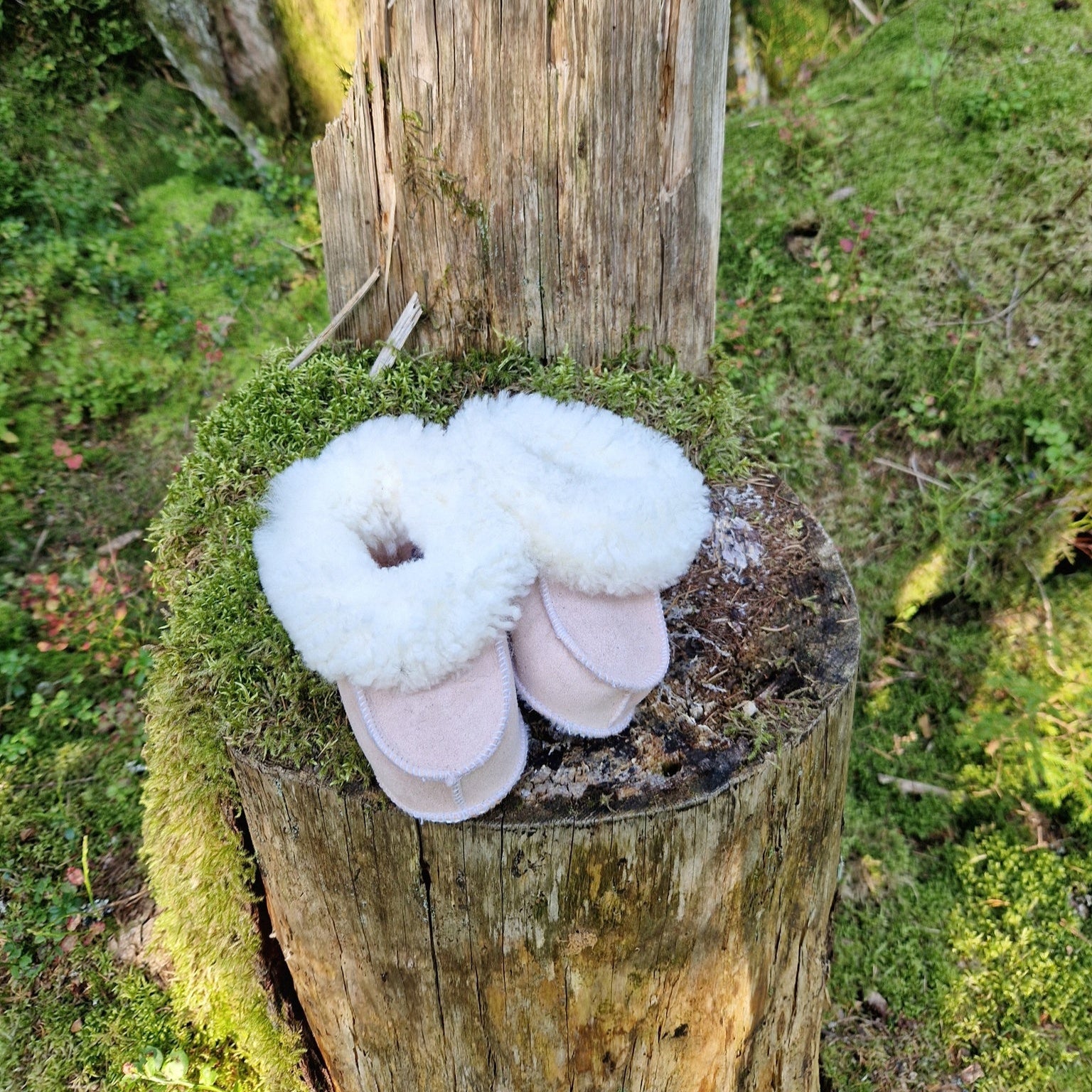 Handgjorda rosa fårskinnstofflor för barn från Jakobs Handmade, placerade på en mossig stubbe i skogen, med vit mjuk fårskinnskrage.