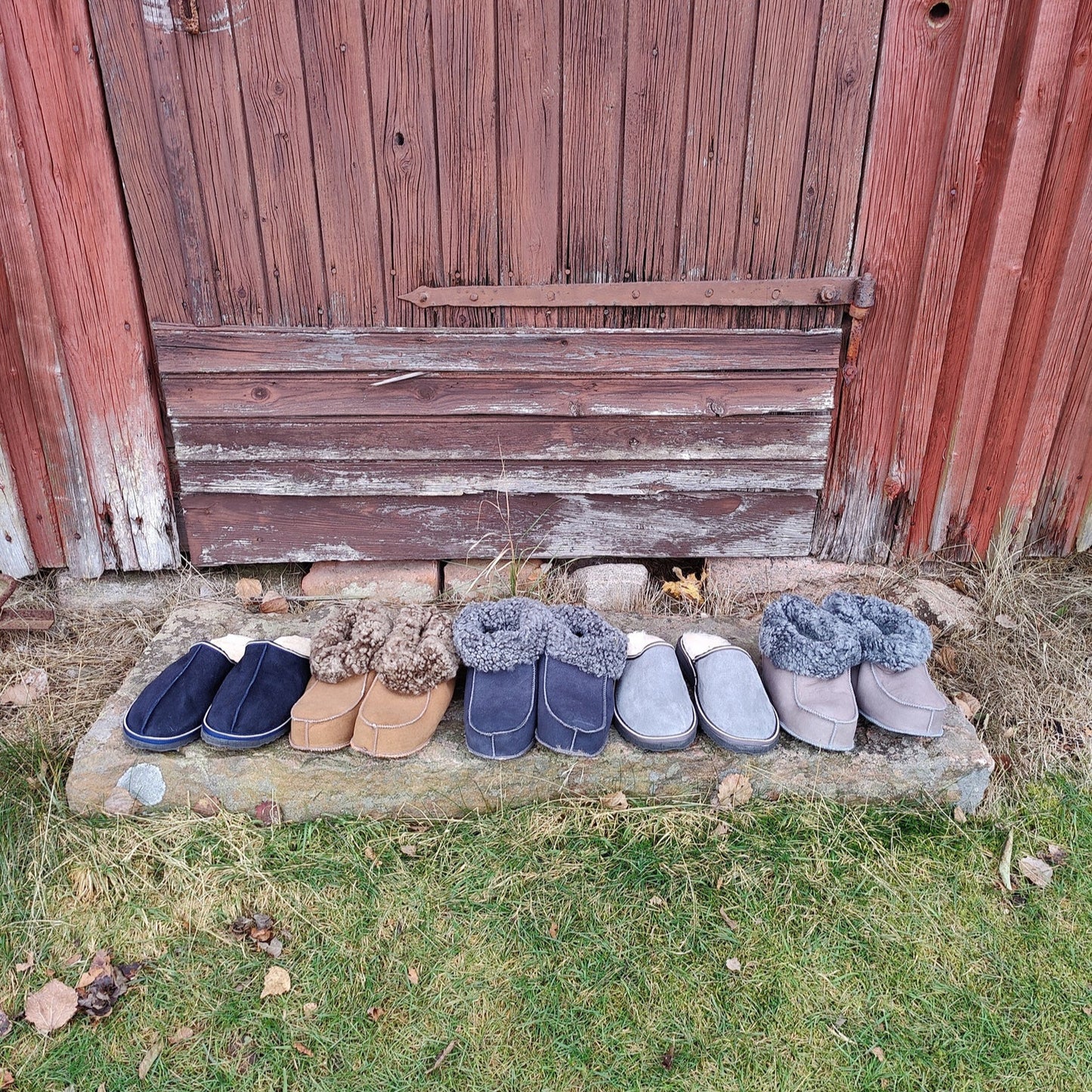 ”Handgjorda fårskinnstofflor i flera färger från Jakobs Handmade, fotograferade utanför en traditionell röd svensk lada. Tillverkade i äkta svenskt fårskinn