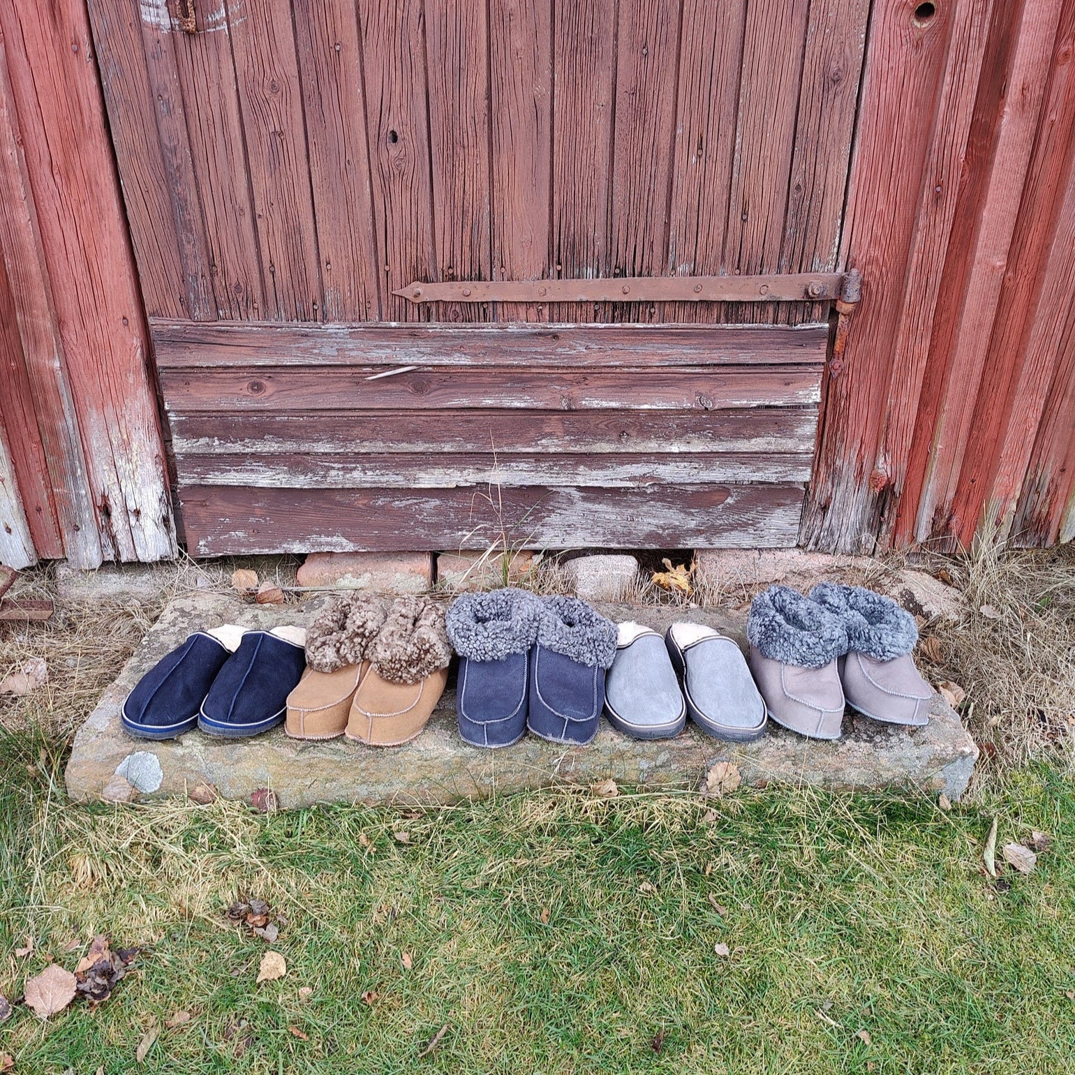 ”Handgjorda fårskinnstofflor i flera färger från Jakobs Handmade, fotograferade utanför en traditionell röd svensk lada. Tillverkade i äkta svenskt fårskinn