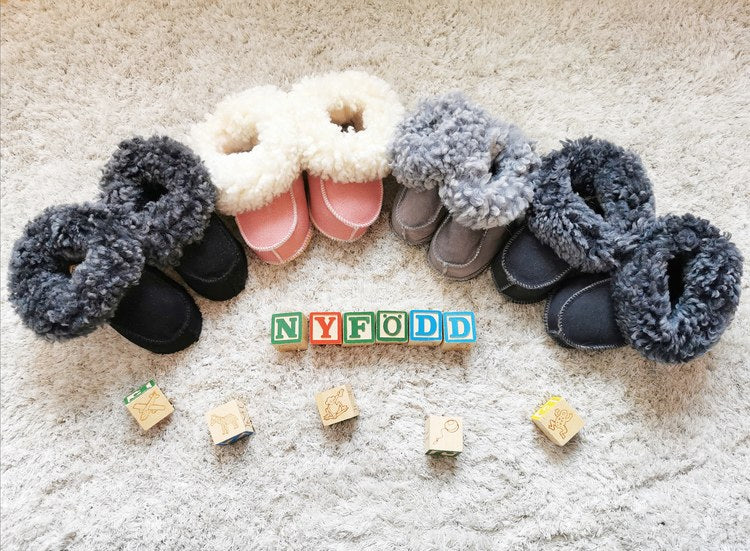 Handgjorda fårskinnstofflor för nyfödda i flera färger, arrangerade i en halvcirkel på en matta med träklossar som stavar "NYFÖDD". Mjuka och värmande tofflor från Jakobs Handmade.