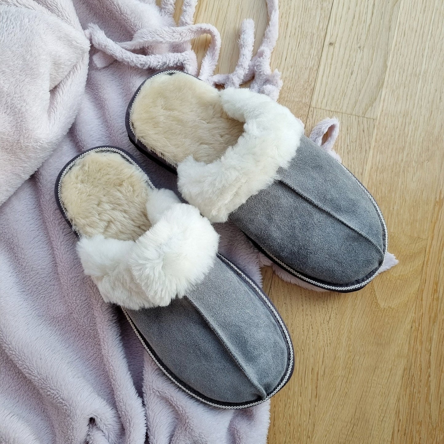 Grå fårskinnstofflor med vitt ullfoder på mjuk rosa pläd, stående på ljust trägolv