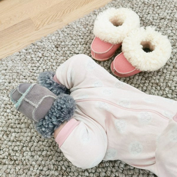 Baby med grå handgjorda fårskinnstofflor från Jakobs Handmade, liggandes på en mjuk matta. Bredvid syns ett par rosa fårskinnstofflor med vit krage.