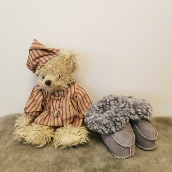 Handgjorda grå fårskinnstofflor för barn från Jakobs Handmade, fotograferade bredvid en nalle i pyjamas. Tofflorna är tillverkade i äkta svenskt fårskinn med mjuk värmande krage.