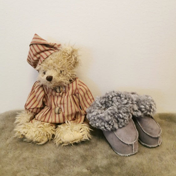 Grå handgjorda fårskinnstofflor för barn från Jakobs Handmade, placerade bredvid en nalle i pyjamas. Tillverkade i äkta svenskt fårskinn med mjuk värmande krage.