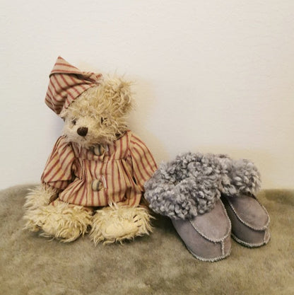 Grå handgjorda fårskinnstofflor för barn från Jakobs Handmade, placerade bredvid en nalle i pyjamas. Tillverkade i äkta svenskt fårskinn med mjuk värmande krage.