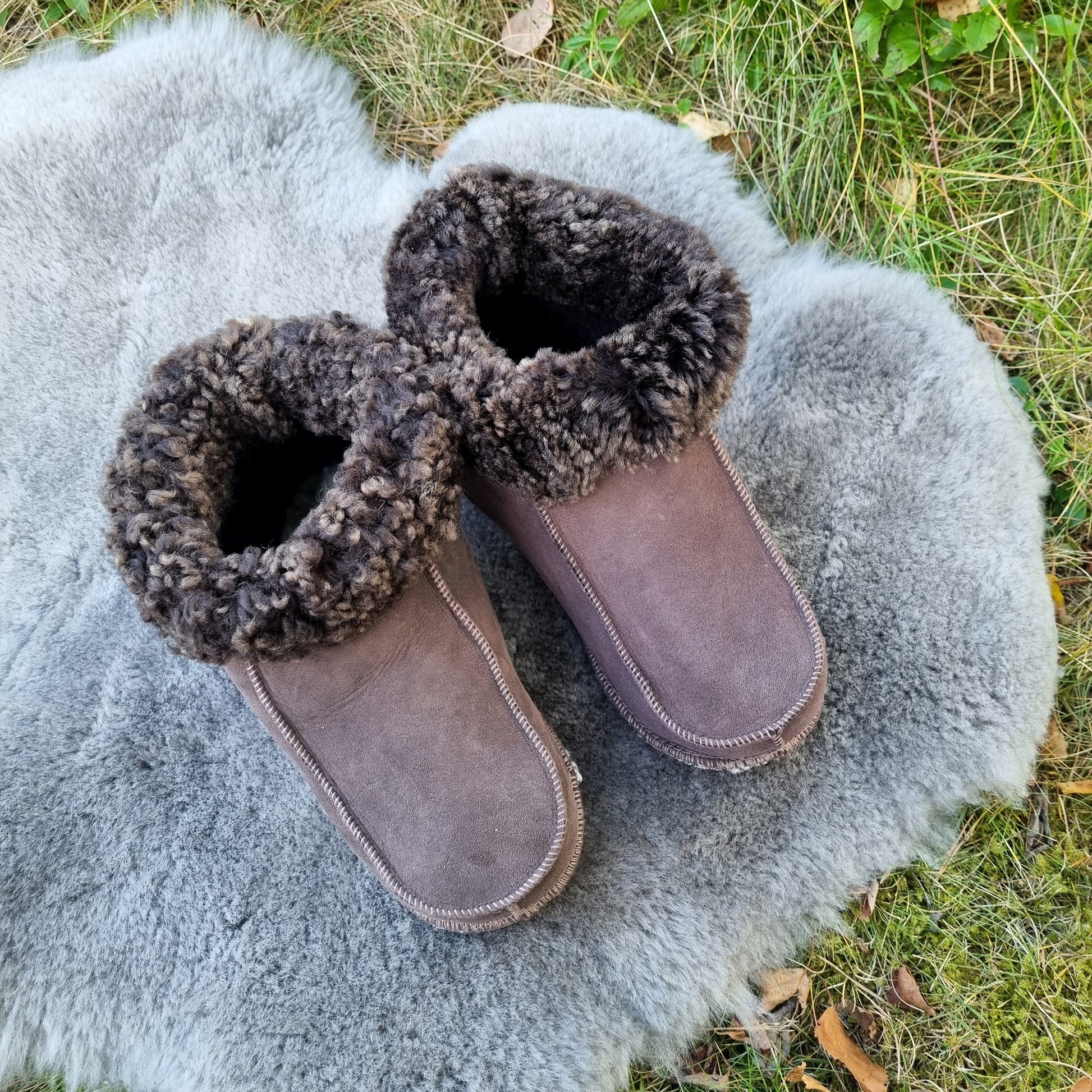 Handgjorda taupe fårskinnstofflor från Jakobs Handmade, placerade på en mjuk grå fårskinnspläd utomhus. Tofflorna är tillverkade i äkta svenskt fårskinn med mörkbrun krage.