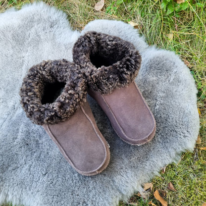 Handgjorda taupe fårskinnstofflor från Jakobs Handmade, placerade på en mjuk grå fårskinnspläd utomhus. Tofflorna är tillverkade i äkta svenskt fårskinn med mörkbrun krage.
