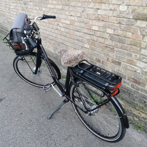 Svart elcykel med ljusbrunt fårskinnsadelskydd och cykelkorg mot tegelvägg