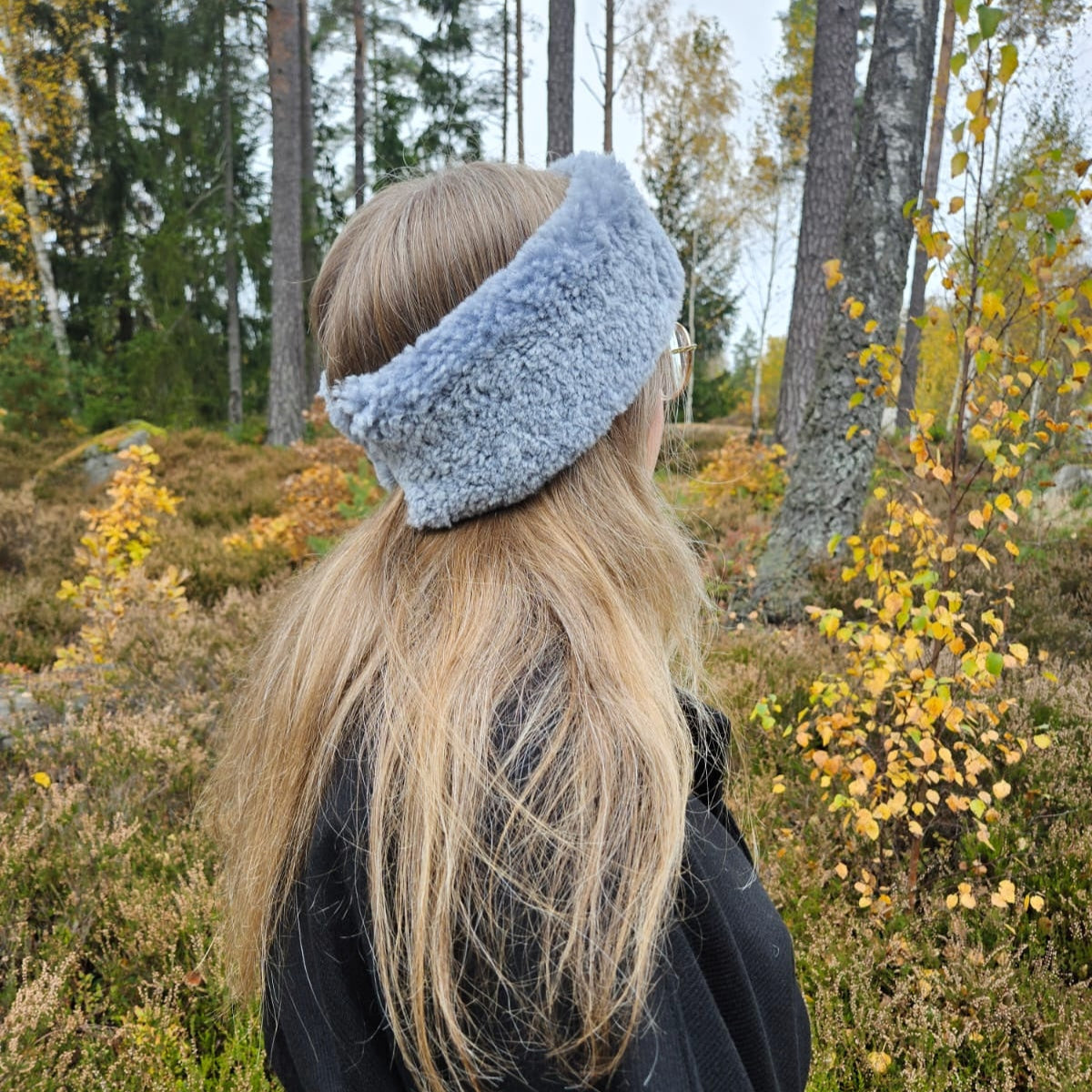 Kvinna med långt blont hår bär ett ljusgrått fårskinnspannband utomhus bland höstlöv och träd.