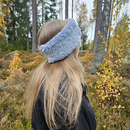 Kvinna med långt blont hår bär ett ljusgrått fårskinnspannband utomhus bland höstlöv och träd.