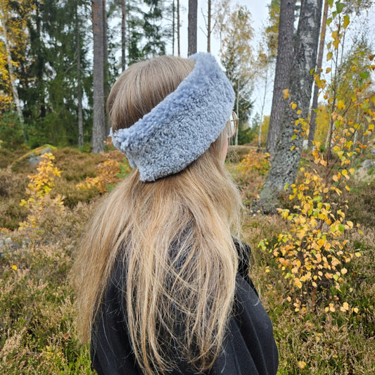 Kvinna med långt blont hår bär ett ljusgrått fårskinnspannband utomhus bland höstlöv och träd.
