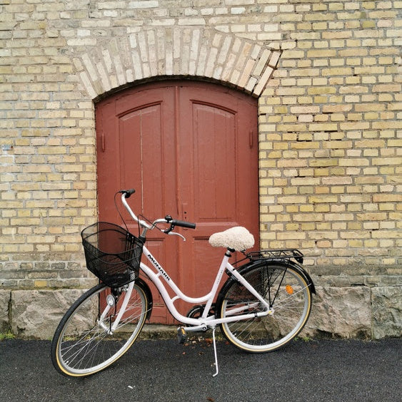 Ljusrosa damcykel med ljust fårskinnsadelskydd och svart cykelkorg parkerad framför en röd trädörr och gul tegelvägg.