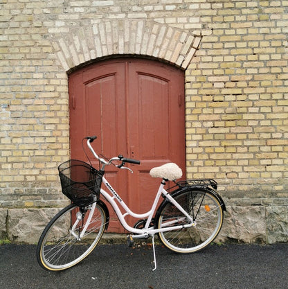 Ljusrosa damcykel med ljust fårskinnsadelskydd och svart cykelkorg parkerad framför en röd trädörr och gul tegelvägg.