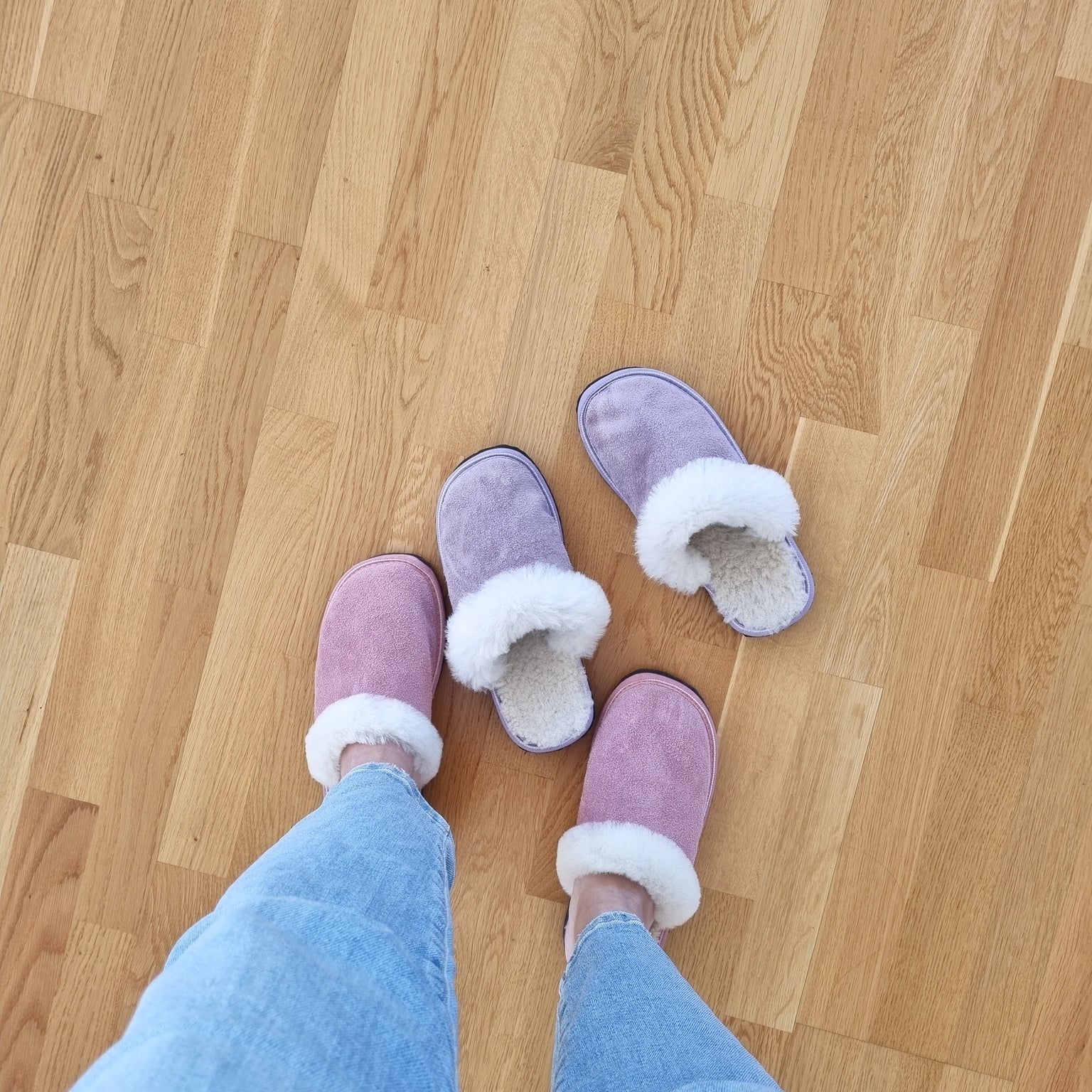 Rosa och lila fårskinnstofflor med vitt ullfoder, en person i jeans bär ett par medan det andra paret står på trägolv, varma och bekväma innetofflor.