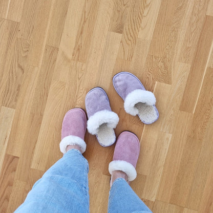 Rosa och lila fårskinnstofflor med vitt ullfoder, en person i jeans bär ett par medan det andra paret står på trägolv, varma och bekväma innetofflor.