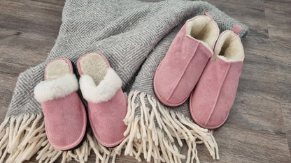 Två par rosa ullstofflor från Jakobs Handmade – Elpida slip-in tofflor med vit fårskinnskant och Kosmos ankelhöga tofflor, placerade på en grå filt med fransar för en varm och hemtrevlig känsla.