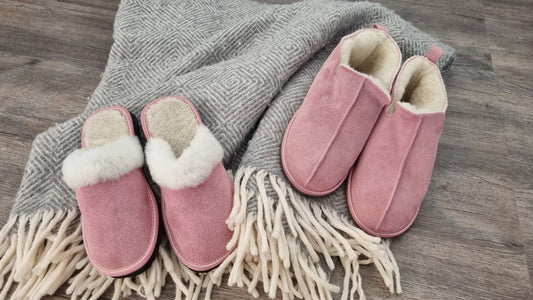 Två par rosa ullstofflor från Jakobs Handmade – Elpida slip-in tofflor med vit fårskinnskant och Kosmos ankelhöga tofflor, placerade på en grå filt med fransar för en varm och hemtrevlig känsla.