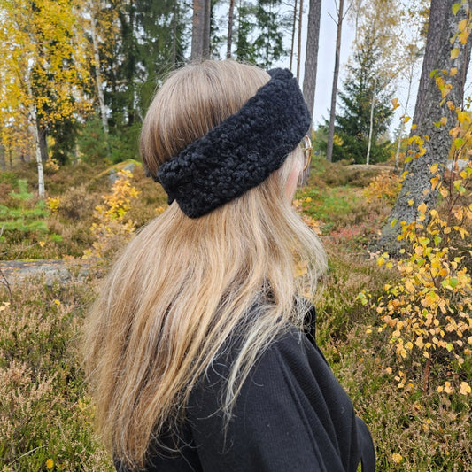 Person med långt blont hår bär ett svart fårskinnspannband i höstig skogsmiljö.