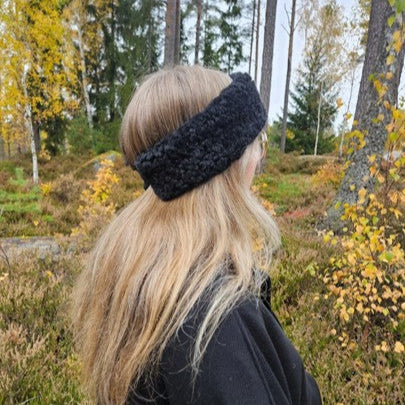 Person med långt blont hår bär ett svart fårskinnspannband i höstig skogsmiljö.